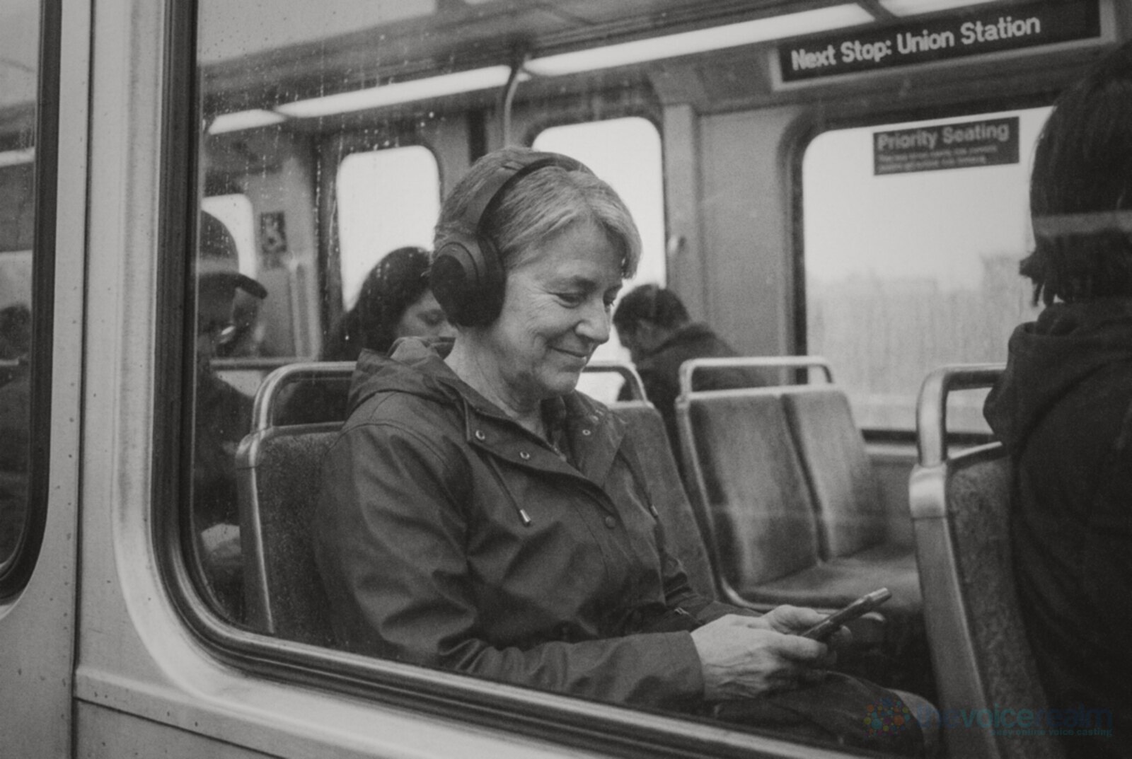 Black and white photo of a woman in her fifties with grey hair and a raincoat on a commuter train, smiling to herself as she listens on over-ear headphones © The Voice Realm