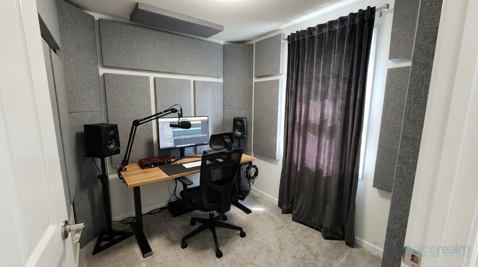 A spare room set up only as a home recording studio, acoustic panels on the walls, heavy curtains, a desk with a monitor, speakers and a microphone on a boom arm © The Voice Realm
