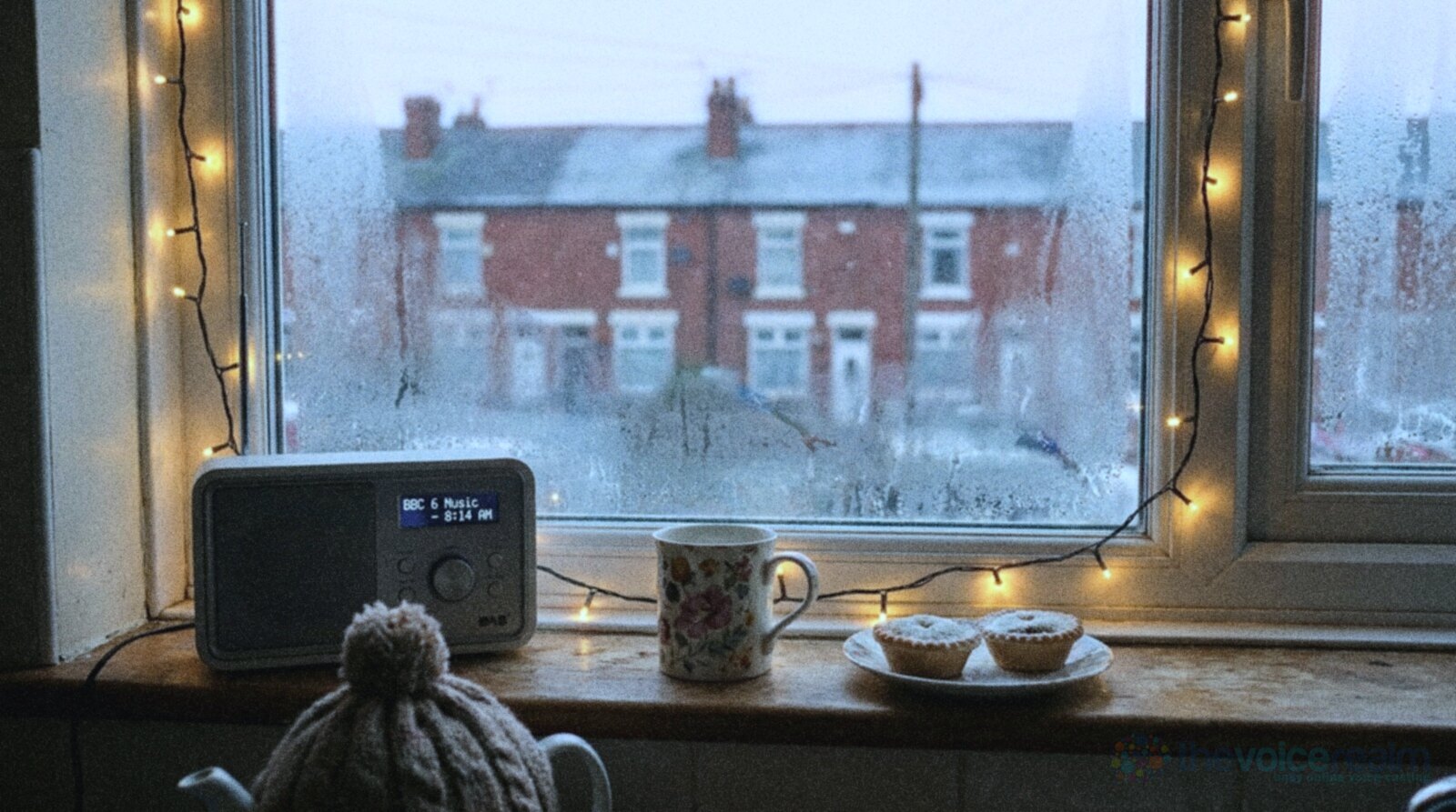 A small DAB radio, a floral mug of tea and two mince pies on the windowsill of a British terraced house kitchen on a frosty morning, fairy lights round the steamy window and red brick houses outside © The Voice Realm