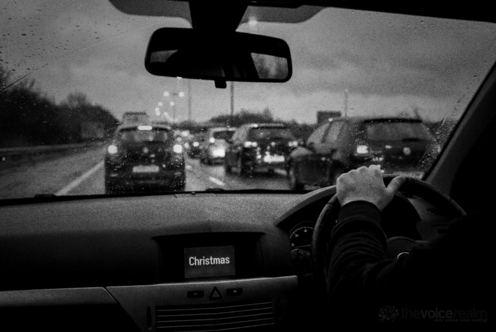 A driver's view of slow traffic on a wet British motorway at dusk, rain on the windscreen and the car radio screen glowing with the word Christmas © The Voice Realm