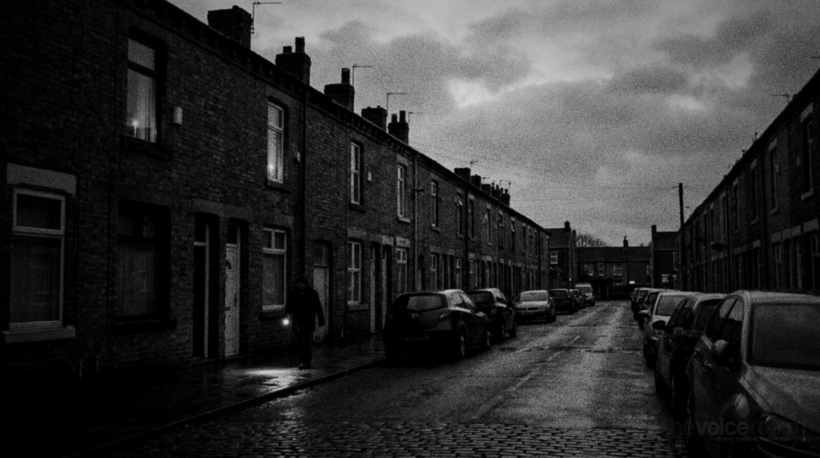 A row of red brick terraced houses on a cobbled street at dusk during a power cut, windows dark except for two candles, a lone person walking along the pavement with a torch © The Voice Realm