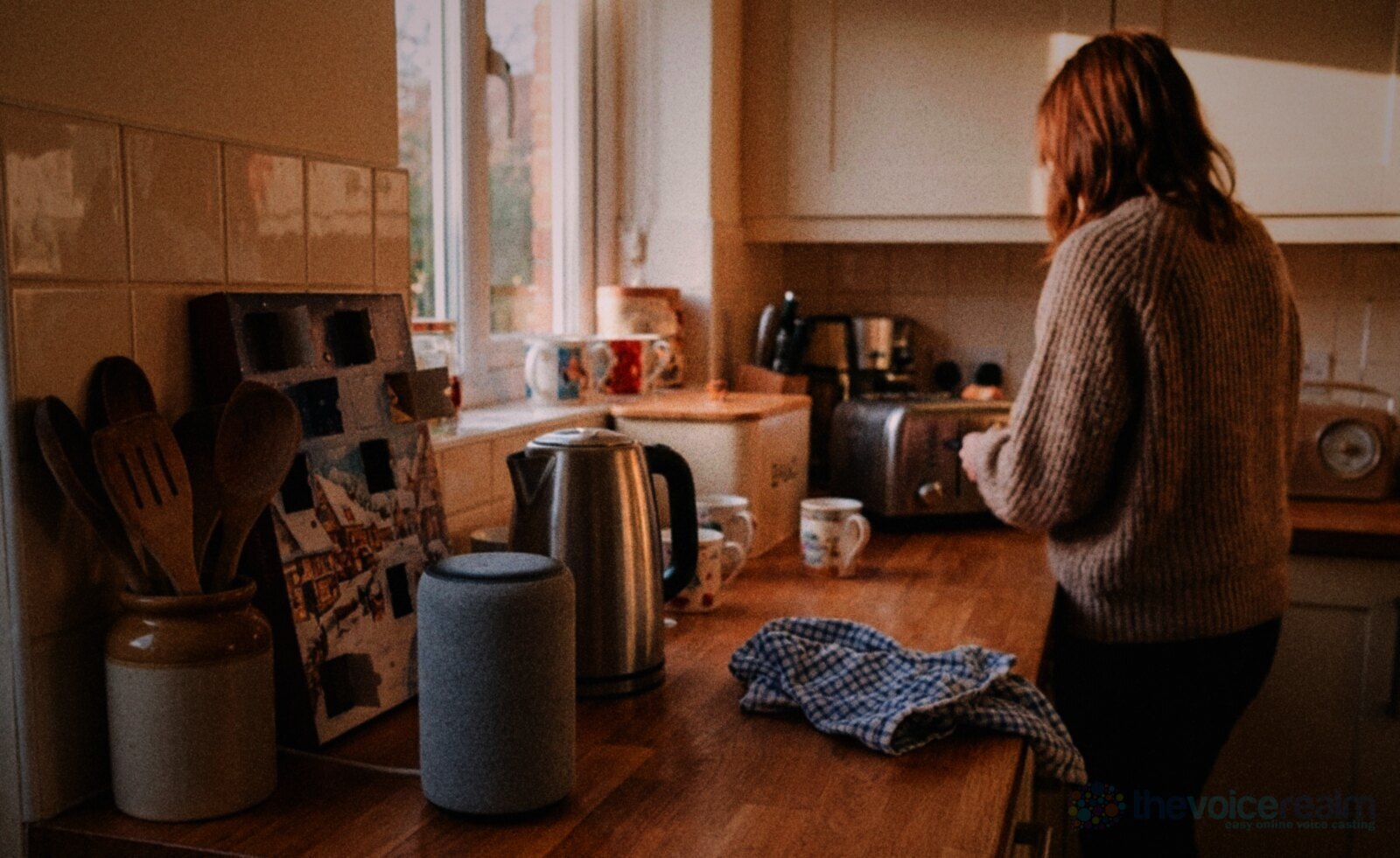 A fabric smart speaker beside a kettle and an advent calendar on a wooden kitchen worktop while a woman in a jumper makes toast in the background © The Voice Realm