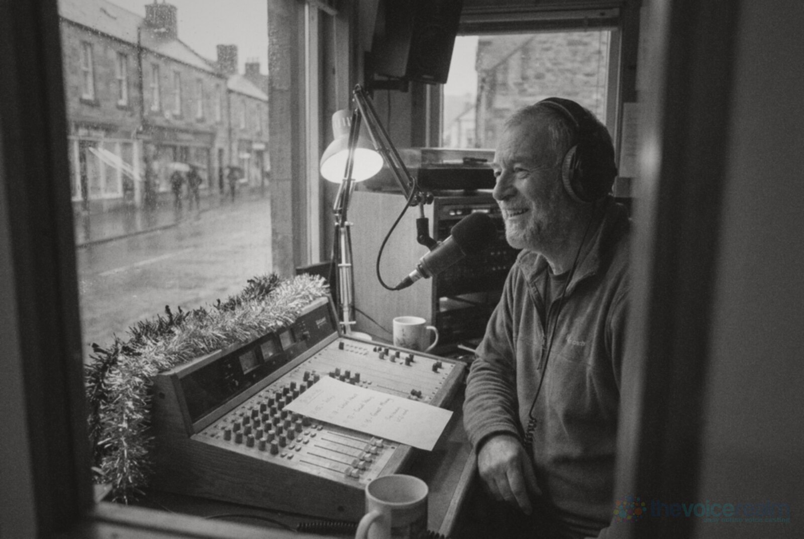 A smiling older presenter in headphones talks into a microphone at a small community radio studio desk trimmed with tinsel, a wet Scottish high street through the window © The Voice Realm