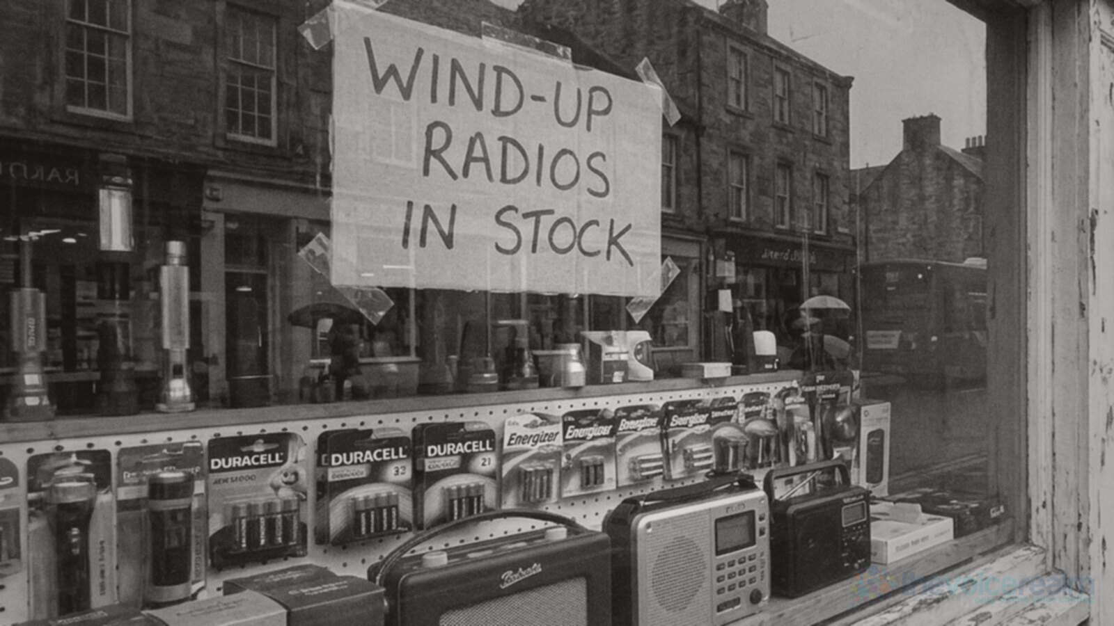 A handwritten sign reading WIND-UP RADIOS IN STOCK taped inside the window of an old hardware shop on a rainy British high street, with torches, batteries and radios on the shelf © The Voice Realm