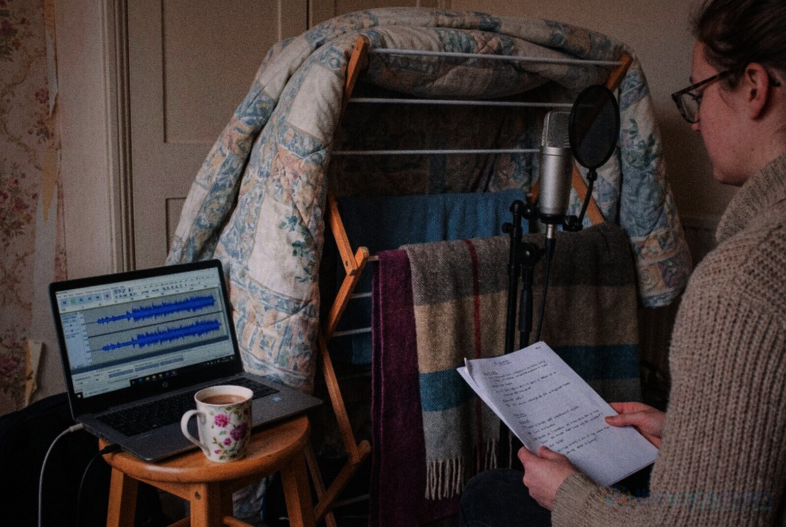 A home voice recording corner in a British terraced house: a duvet and blankets hung over a clothes airer behind a microphone, a laptop showing a waveform on a stool and a mug of tea, a narrator holding printed pages © The Voice Realm