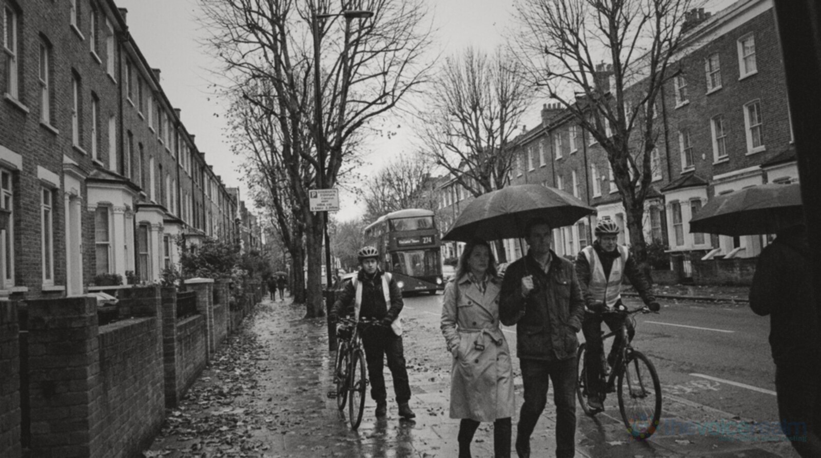 Black and white street photo of a tree-lined road of Victorian terraces in Camden, London on a wet autumn morning, people under umbrellas, two cyclists and a double-decker bus in the distance © The Voice Realm