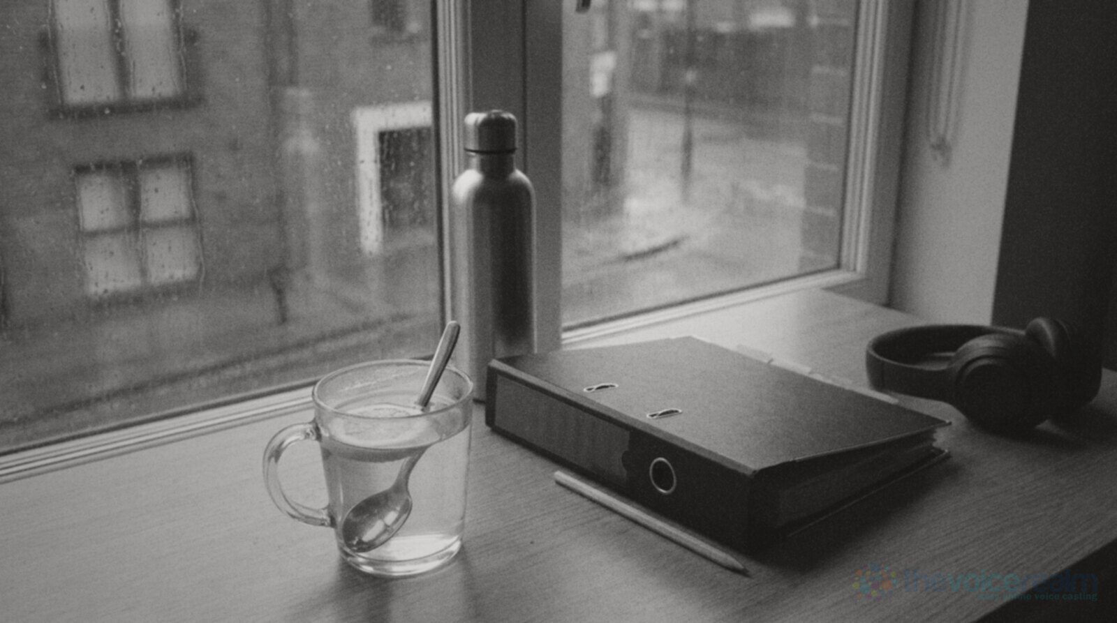 Black and white photo of a desk by a rainy window in a London flat with a glass mug of hot water, lemon and honey, a water bottle, a closed ring binder with coloured tabs, a pencil and headphones © The Voice Realm