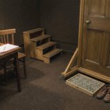 The spot effects corner of a radio drama studio with nobody in it: a wooden door standing in its own frame on castors, a tray of gravel and a pair of brogues on the carpet, practice stairs, and a kitchen table with a brown teapot, cups and a kettle under a lamp, in cool grainy film colours © The Voice Realm