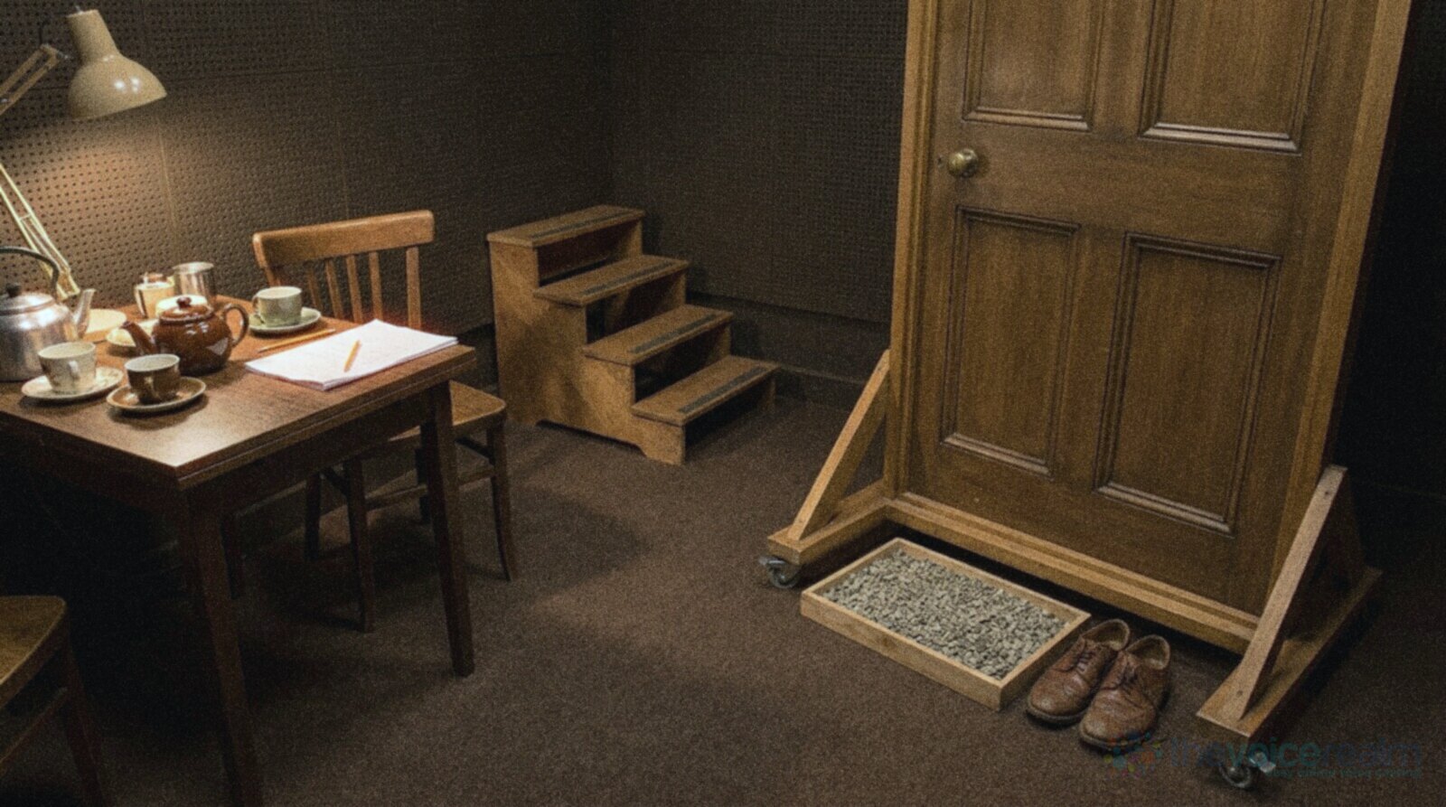 The spot effects corner of a radio drama studio with nobody in it: a wooden door standing in its own frame on castors, a tray of gravel and a pair of brogues on the carpet, practice stairs, and a kitchen table with a brown teapot, cups and a kettle under a lamp, in cool grainy film colours © The Voice Realm