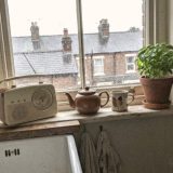 A rainy morning in the kitchen of a Victorian terraced house, a cream portable radio with a round tuning dial on the windowsill beside a brown teapot, a mug and a pot of basil, rain on the sash window and wet slate roofs and chimney pots across the street, in bright phone snapshot colours © The Voice Realm