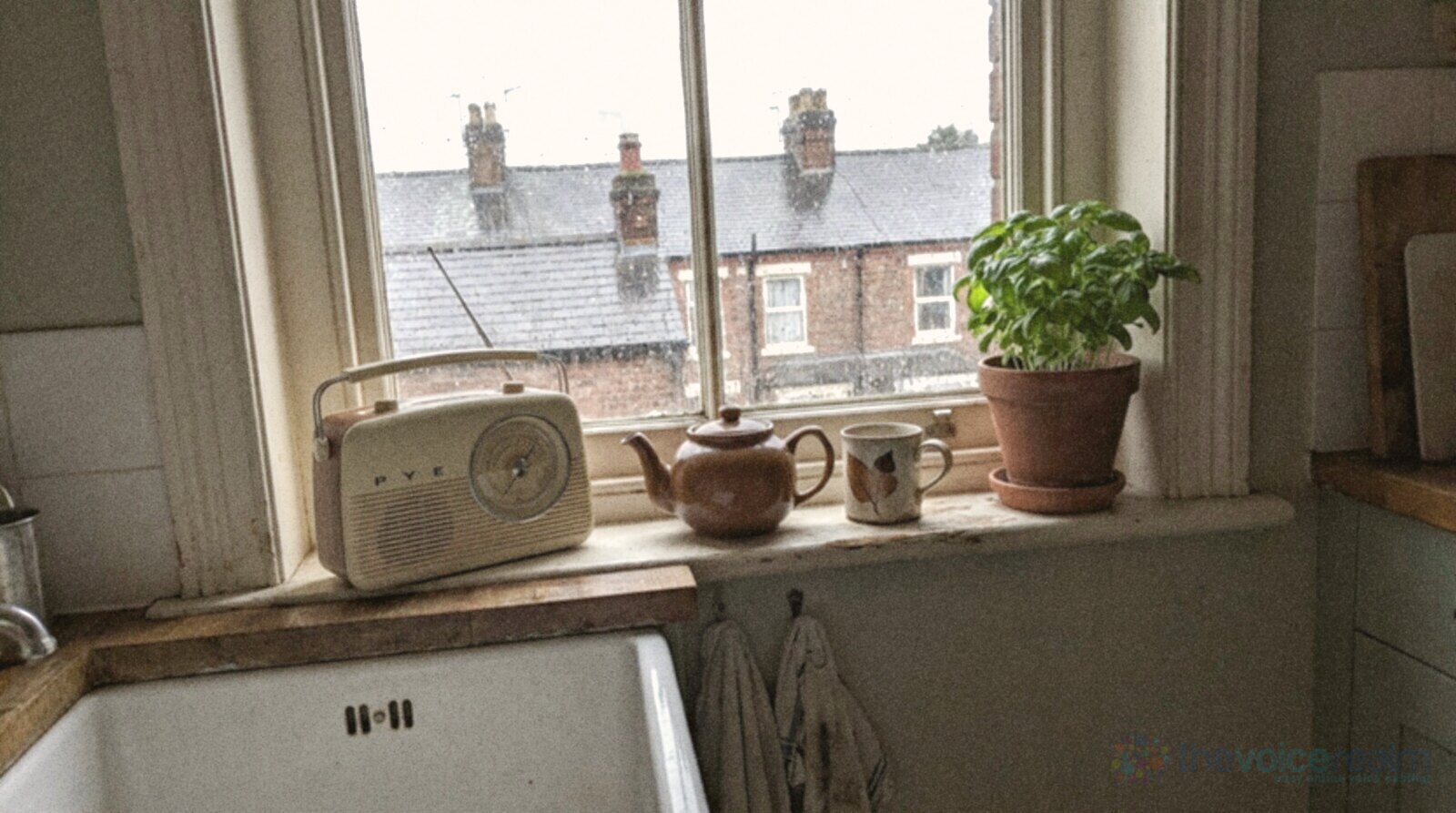 A rainy morning in the kitchen of a Victorian terraced house, a cream portable radio with a round tuning dial on the windowsill beside a brown teapot, a mug and a pot of basil, rain on the sash window and wet slate roofs and chimney pots across the street, in bright phone snapshot colours © The Voice Realm