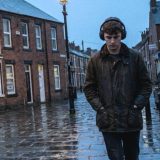 A young man in a waxed jacket and over-ear headphones walking along a wet street of red brick terraced houses in a northern English town at dusk, a lit fish and chip shop and a black cab behind him © The Voice Realm