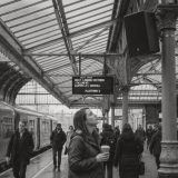 Black and white photo of commuters on a covered Victorian railway platform in Britain, a woman in a raincoat holding a coffee looks up at a loudspeaker on an iron pillar beside a departure board © The Voice Realm
