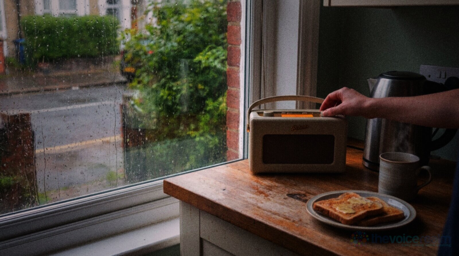 A small cream digital radio on a worn wooden worktop in a terraced house kitchen, a hand reaching for the dial, toast and a mug of tea beside a rain-streaked window © The Voice Realm