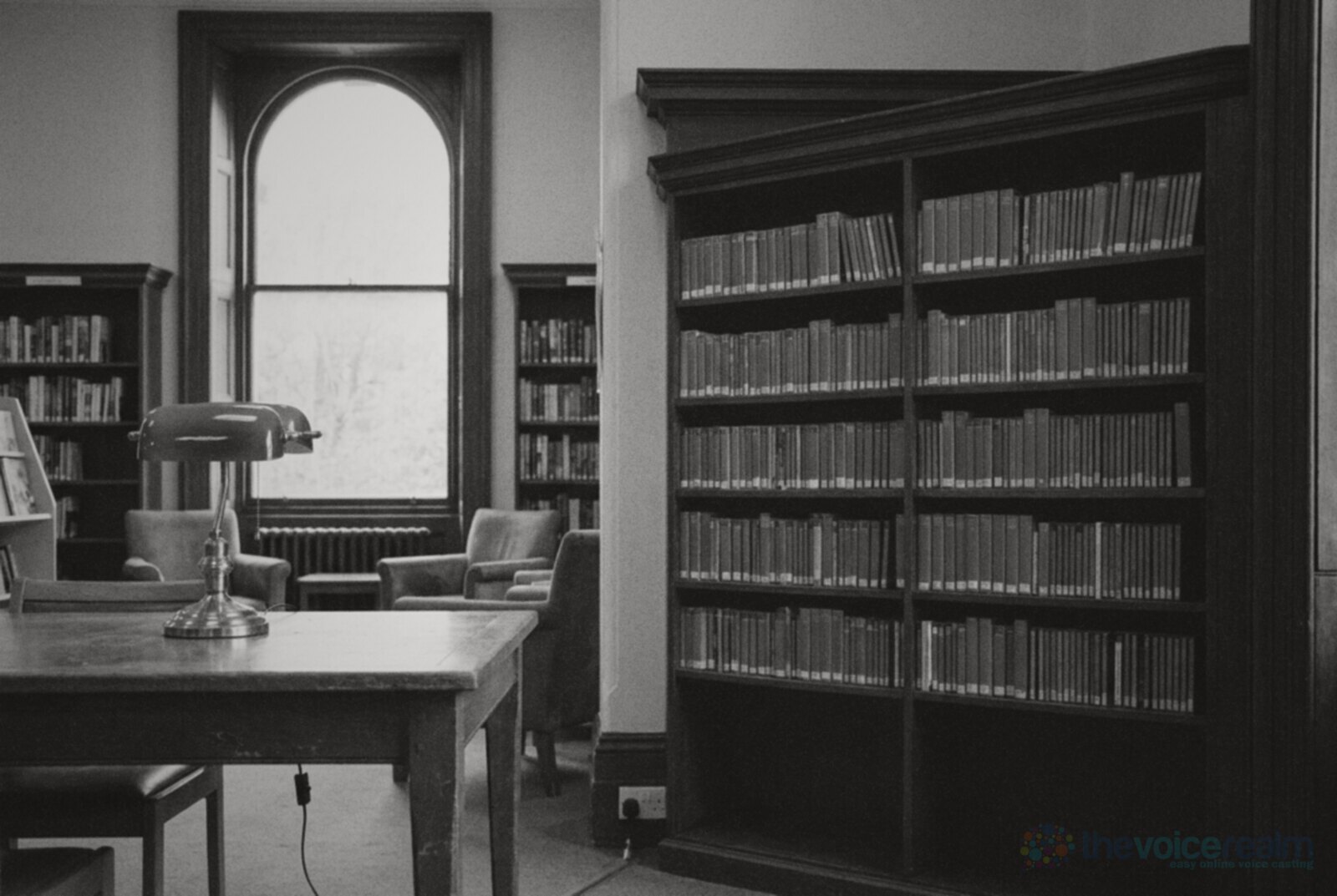 A quiet British public library with tall bookcases, a green-shaded desk lamp on a wooden table and daylight from an arched window © The Voice Realm
