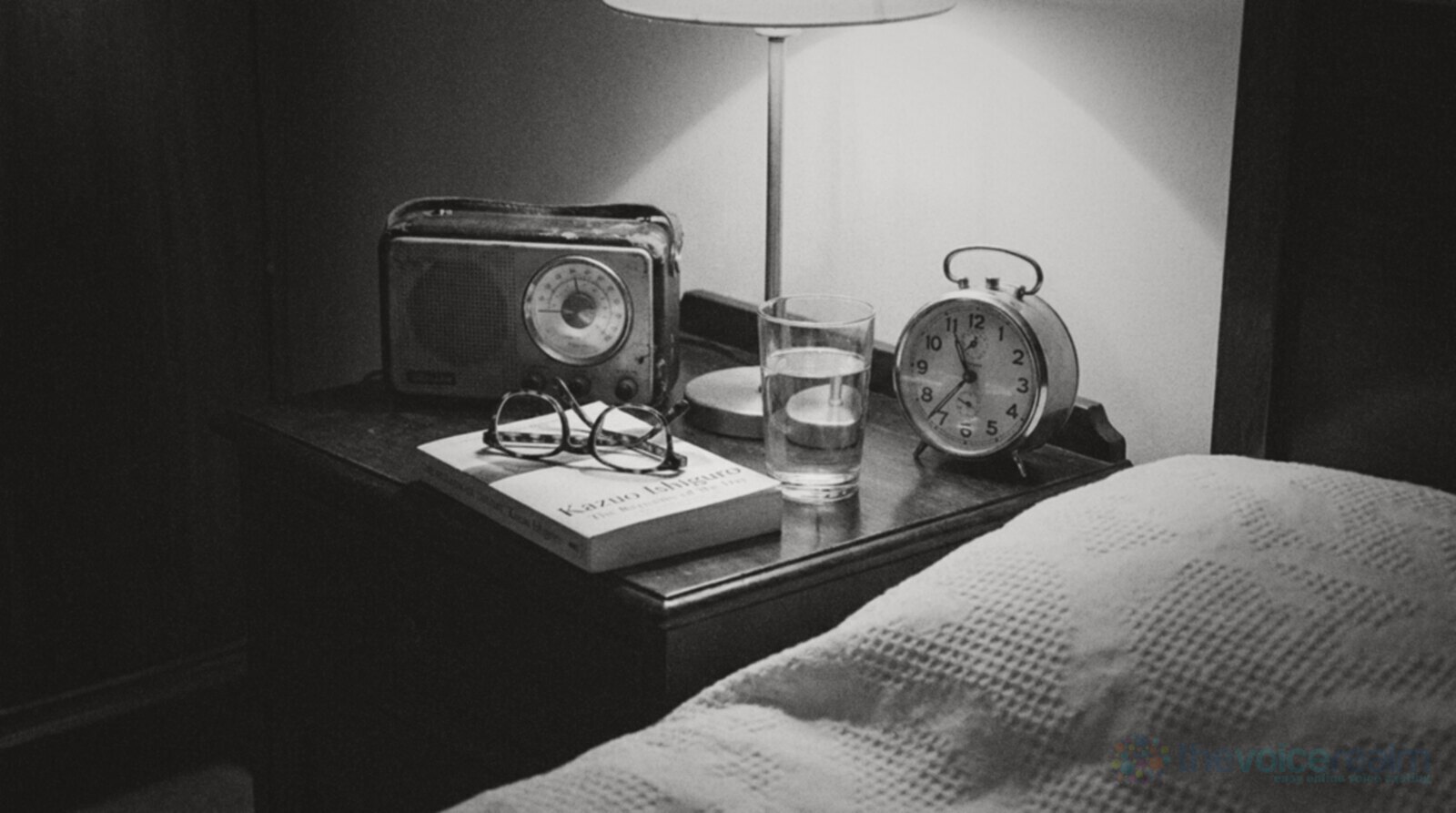 Black and white photo of a bedside table at night by lamp light, an old portable radio with a glowing round dial, reading glasses folded on a paperback, a glass of water and a wind up alarm clock, the edge of a white bedspread in the foreground © The Voice Realm