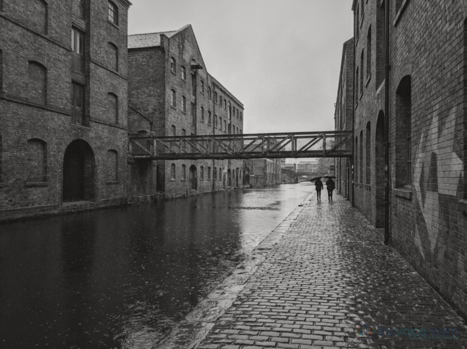 Black and white photo of a canal towpath in Digbeth, Birmingham, on a wet morning, red brick Victorian warehouses on both sides, an iron footbridge over the water, graffiti on a wall and two people walking away under umbrellas © The Voice Realm
