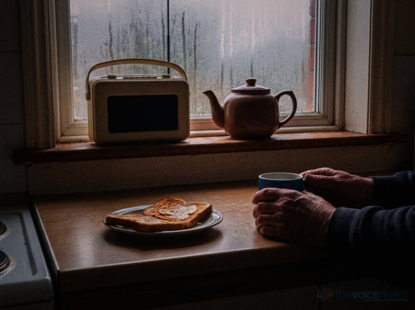 An older man's hands around a blue mug in a small terraced house kitchen on a Sunday morning, a cream portable radio and a brown teapot on the windowsill, buttered toast on a plate and condensation on the window, in punchy film colours © The Voice Realm