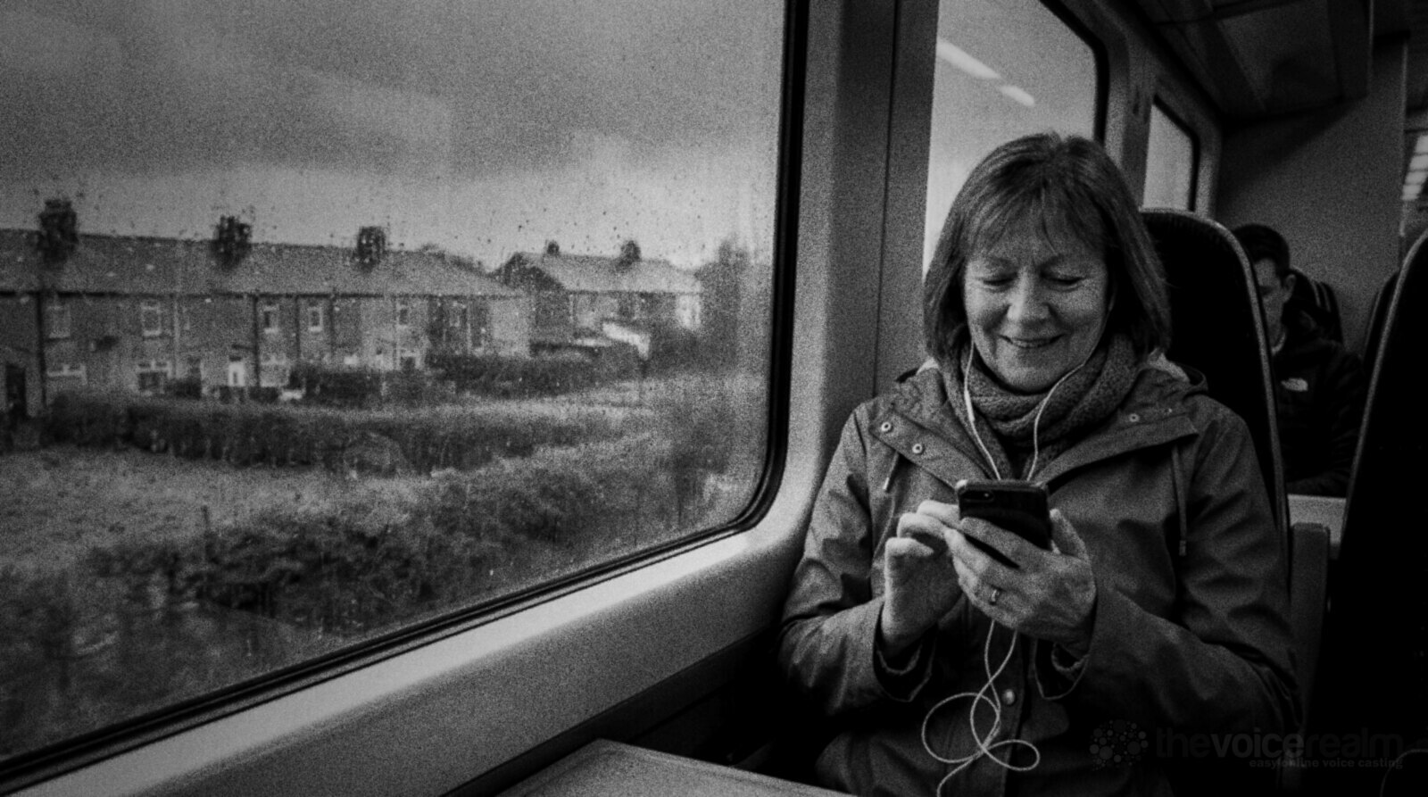 Black and white photo of a woman in a raincoat and scarf on a commuter train, smiling at her phone as she listens through wired earphones, rain on the window and a row of terraced houses outside © The Voice Realm