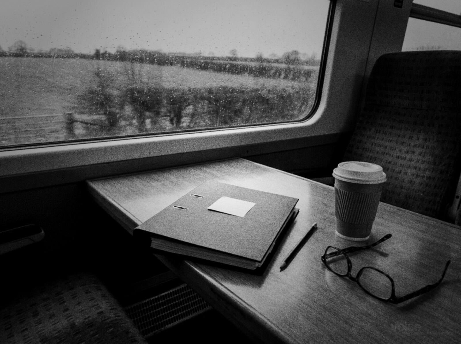 Black and white photo of a table seat on a British train on a rainy day, a closed black ring binder with a sticky note, a pencil, a takeaway coffee and reading glasses by a window streaked with rain over green fields © The Voice Realm