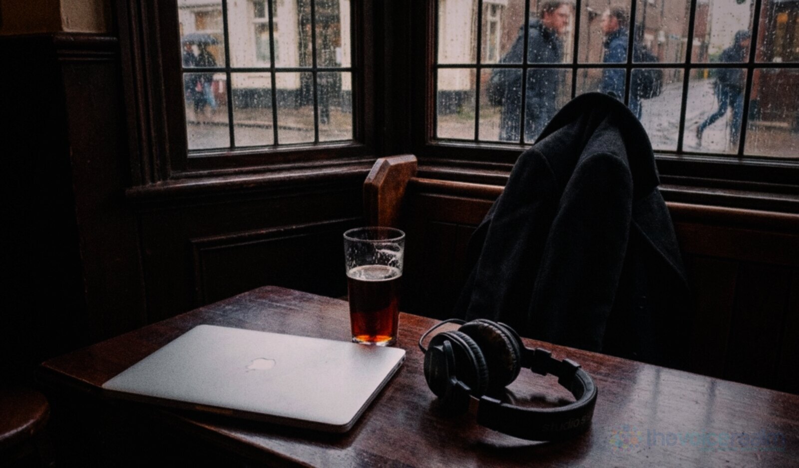 A closed laptop, studio headphones and a half pint of bitter on a dark wooden table in the corner of a traditional British pub, a coat on the settle and rain on the leaded window © The Voice Realm