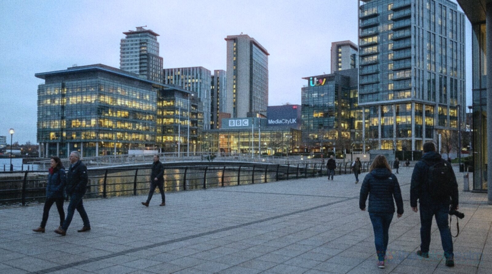MediaCityUK at Salford Quays at dusk, glass office buildings lit up around the piazza and the water, with people walking across the square © The Voice Realm