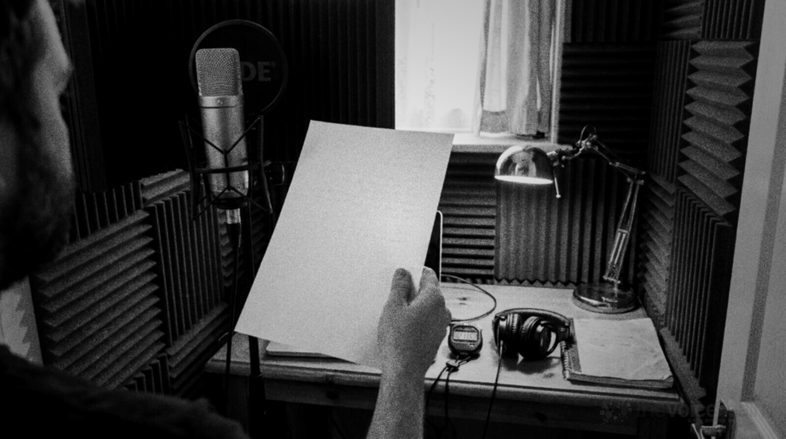 A voiceover artist in a small home booth lined with acoustic foam holds up a sheet of paper beside a condenser microphone, with a stopwatch, headphones and a desk lamp on the desk © The Voice Realm