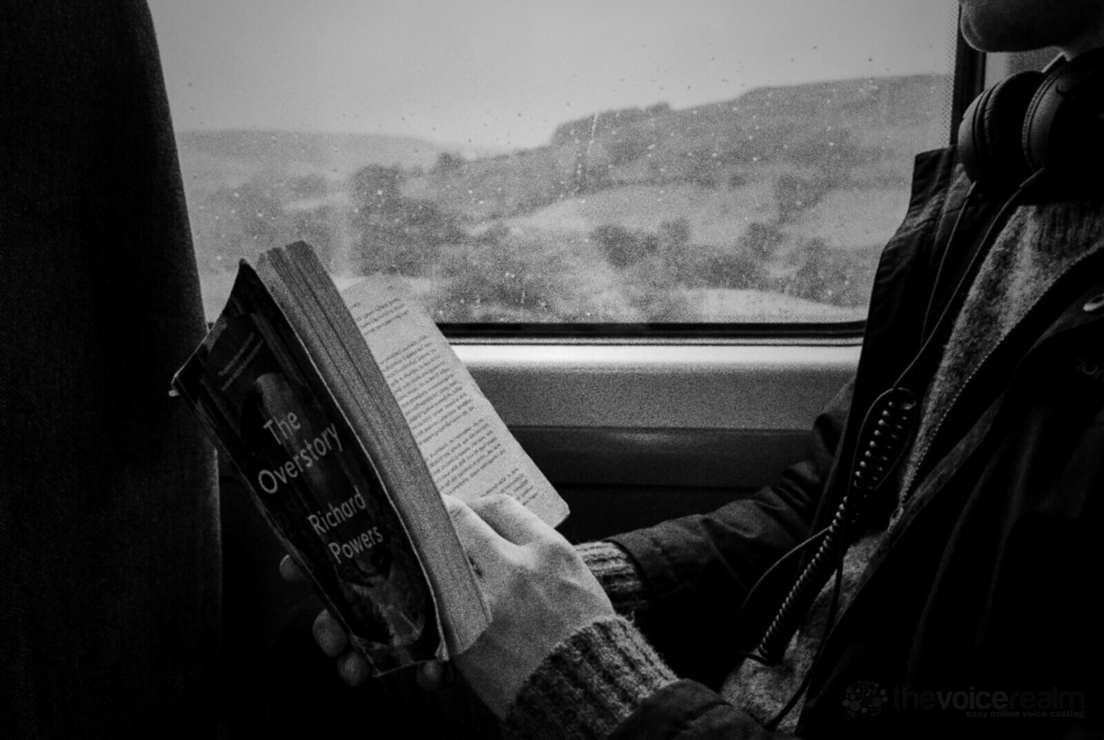 A passenger on a British train holding a well-read paperback, wired headphones around their neck, green rainy hills passing the window © The Voice Realm