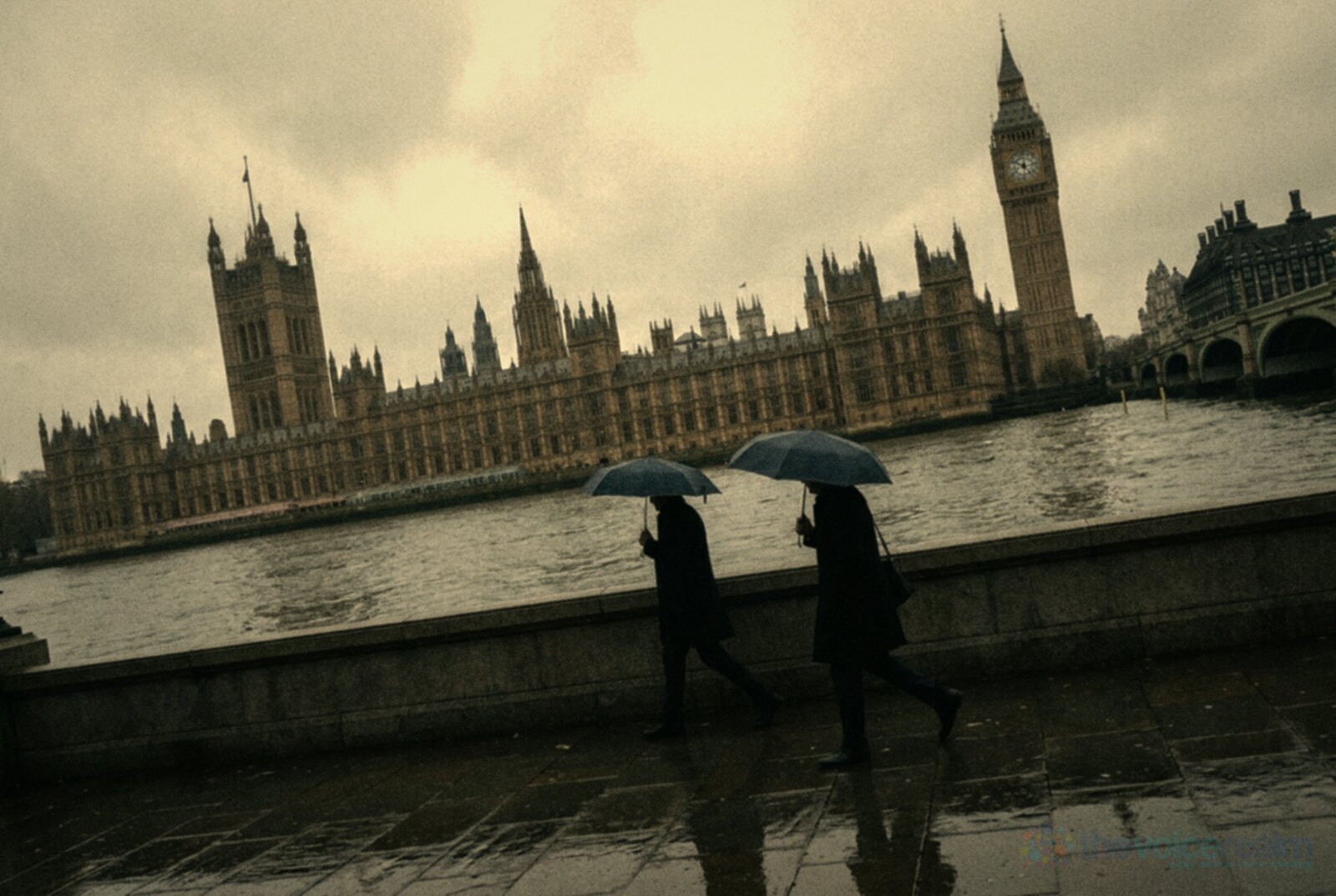 The Palace of Westminster and Elizabeth Tower across the River Thames on an overcast morning, two people with umbrellas walking along the wet embankment © The Voice Realm