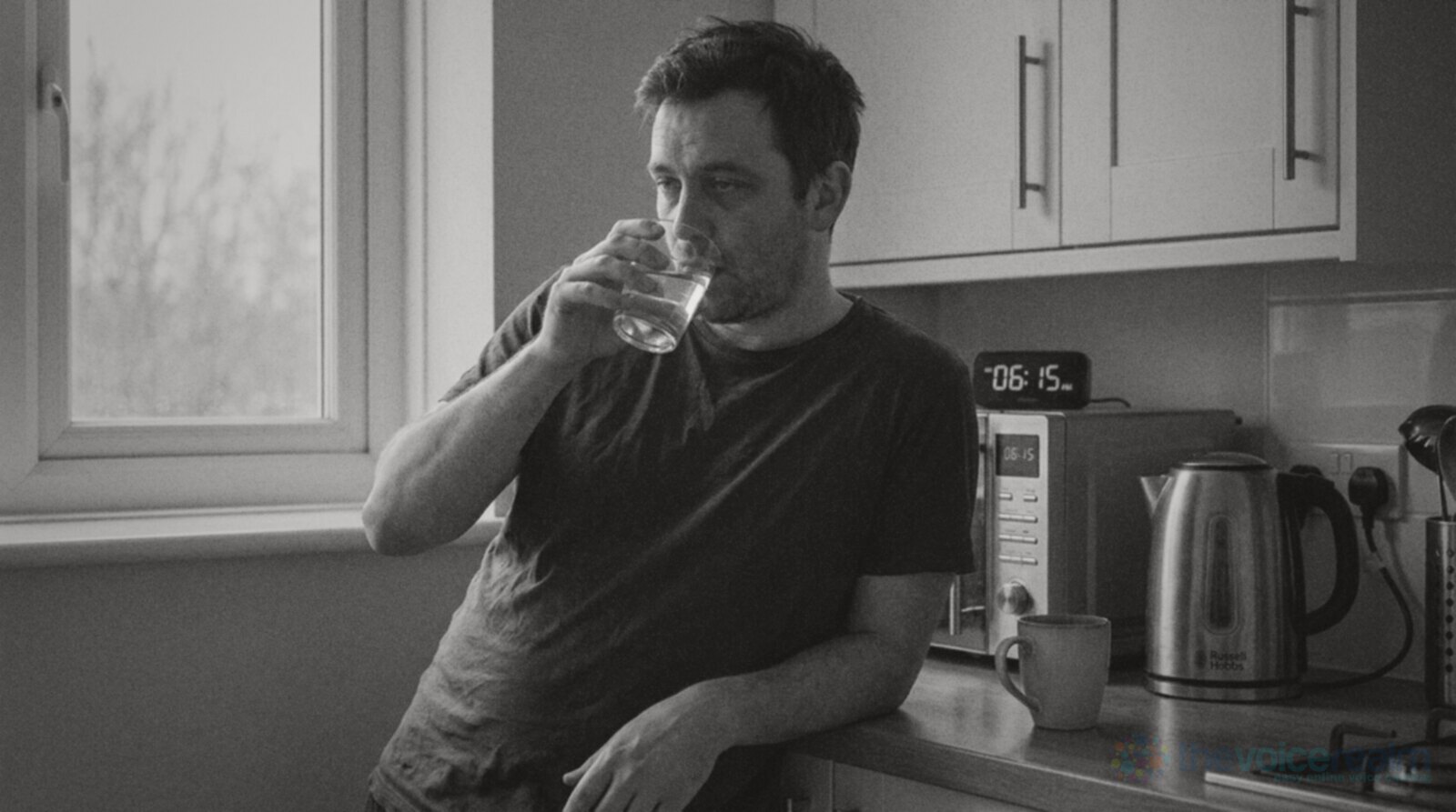 A sleepy man in a dark t-shirt leaning on a kitchen counter at a quarter past six in the morning, drinking a glass of water beside a kettle and a mug © The Voice Realm