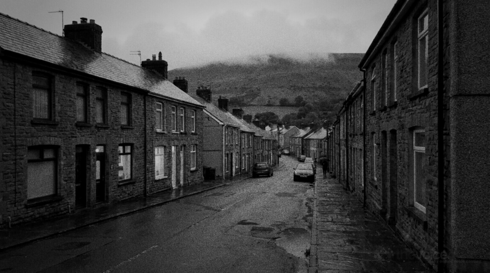 Black and white photo of a terraced street in a Welsh valley village at dusk after rain, two rows of stone houses with wet slate roofs climbing towards a green hillside under low cloud, a few windows lit © The Voice Realm