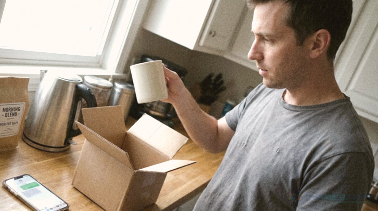 A man in a grey T-shirt lifts a plain white mug out of an opened cardboard delivery box on his kitchen counter and looks it over, a kettle and a phone beside him, in bright phone snapshot colours © The Voice Realm