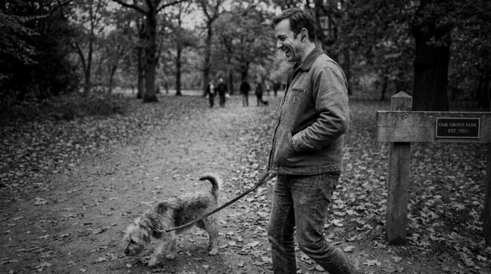 A man in a canvas jacket laughs to himself as he walks a scruffy dog through a leafy autumn park with an earbud in, black and white photograph © The Voice Realm