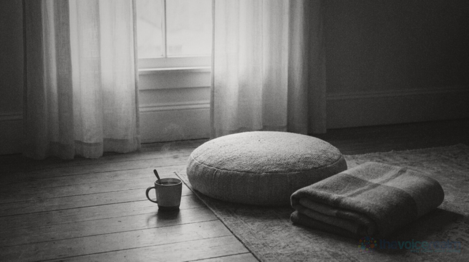 A round woven floor cushion and a folded wool blanket on a wooden floor beside tall sheer curtains at dawn, with a mug of tea on the boards next to them and nobody in the room, black and white photograph © The Voice Realm