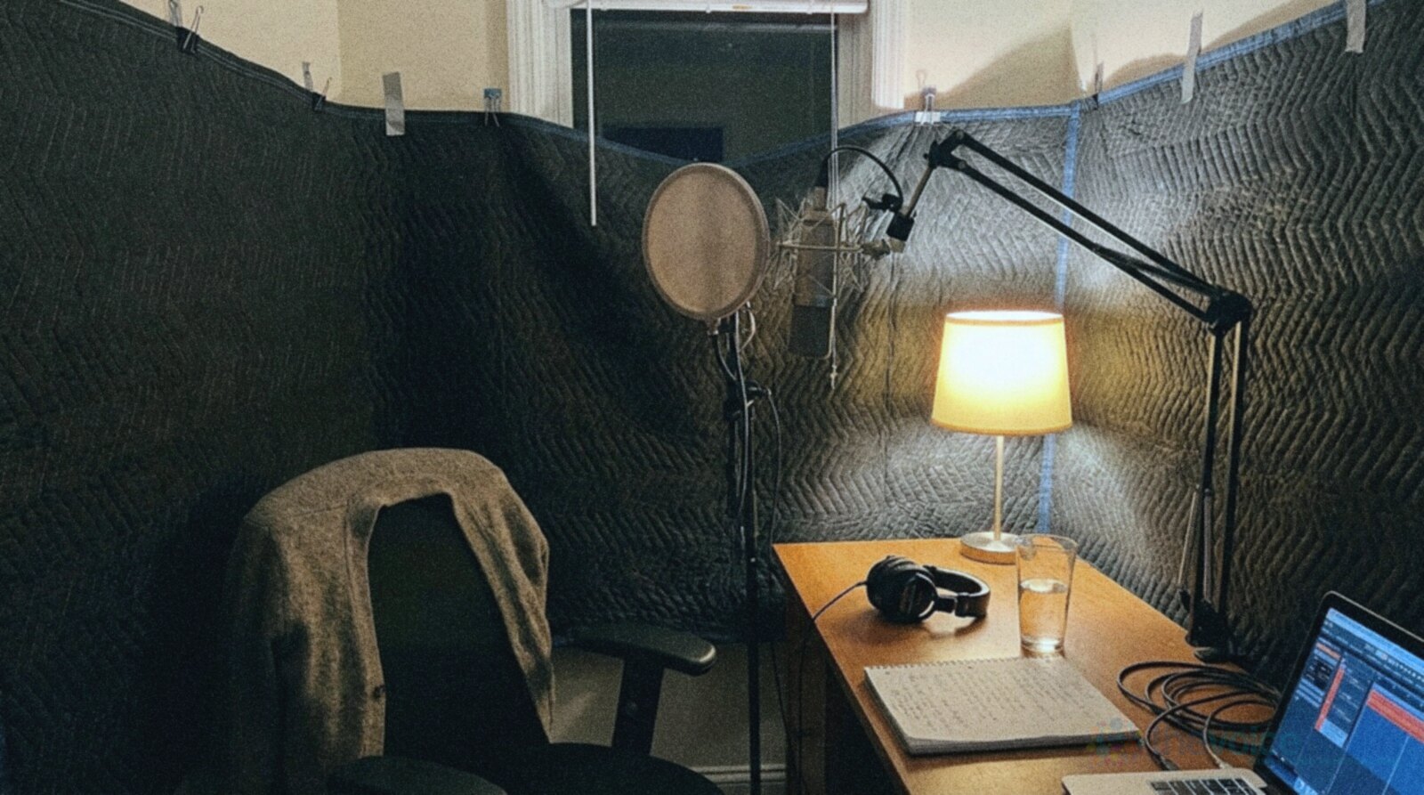 A small home recording corner at night lined with quilted moving blankets clipped to the walls, a microphone with a round pop filter, a dim desk lamp, headphones, a glass of water and a notebook on the desk and a cardigan over the chair © The Voice Realm