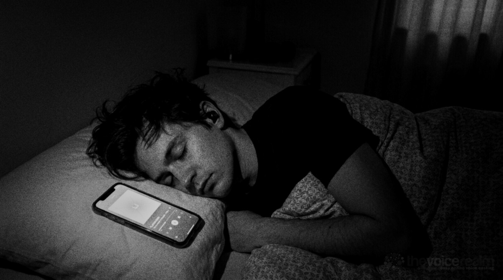 A young man asleep on his side in a dark bedroom with a wireless earbud in, a phone glowing on the pillow beside him showing an audio player, black and white photograph © The Voice Realm