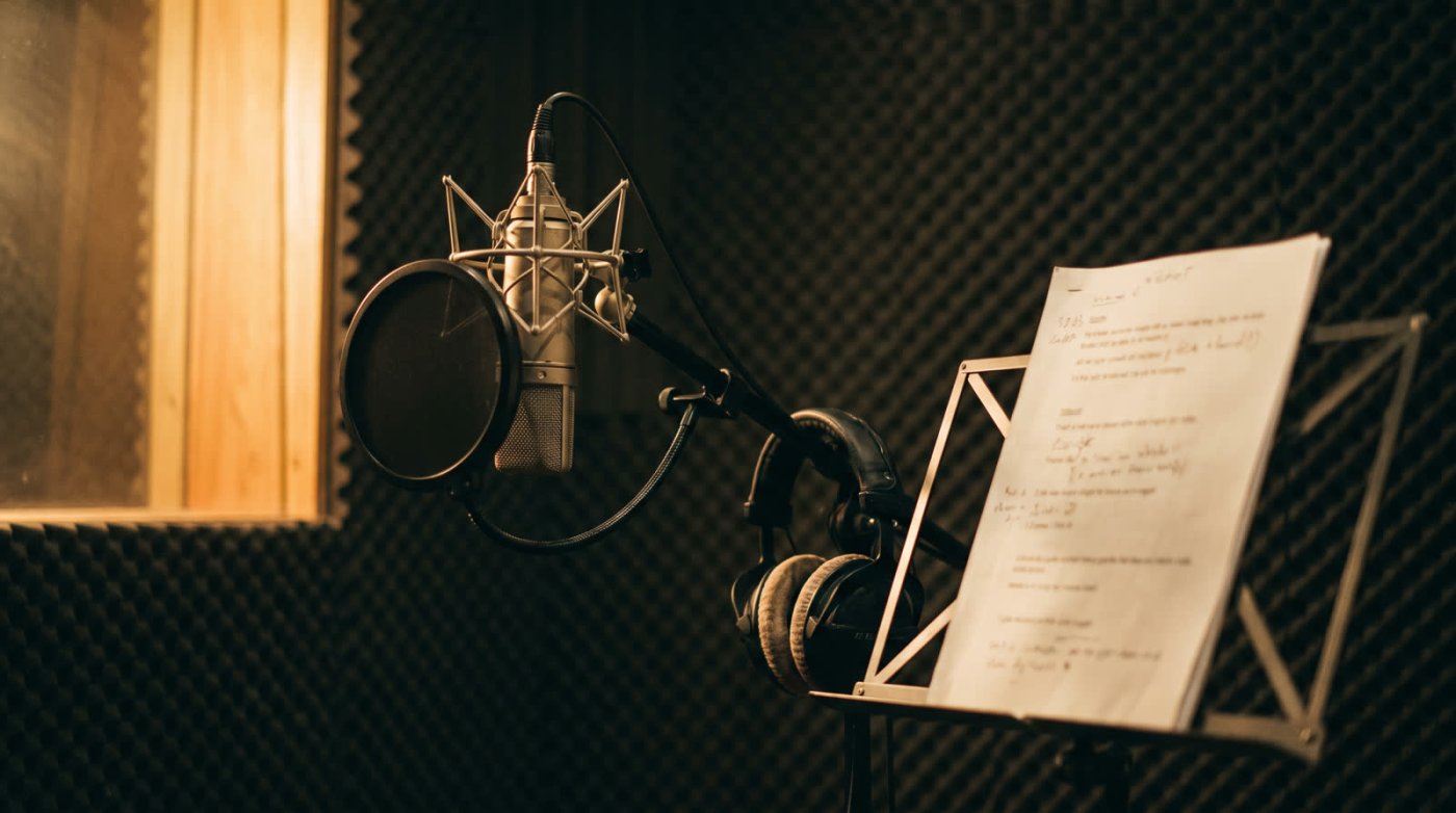 A studio microphone in a shock mount with a pop filter in front of it, headphones and a marked-up script on a music stand